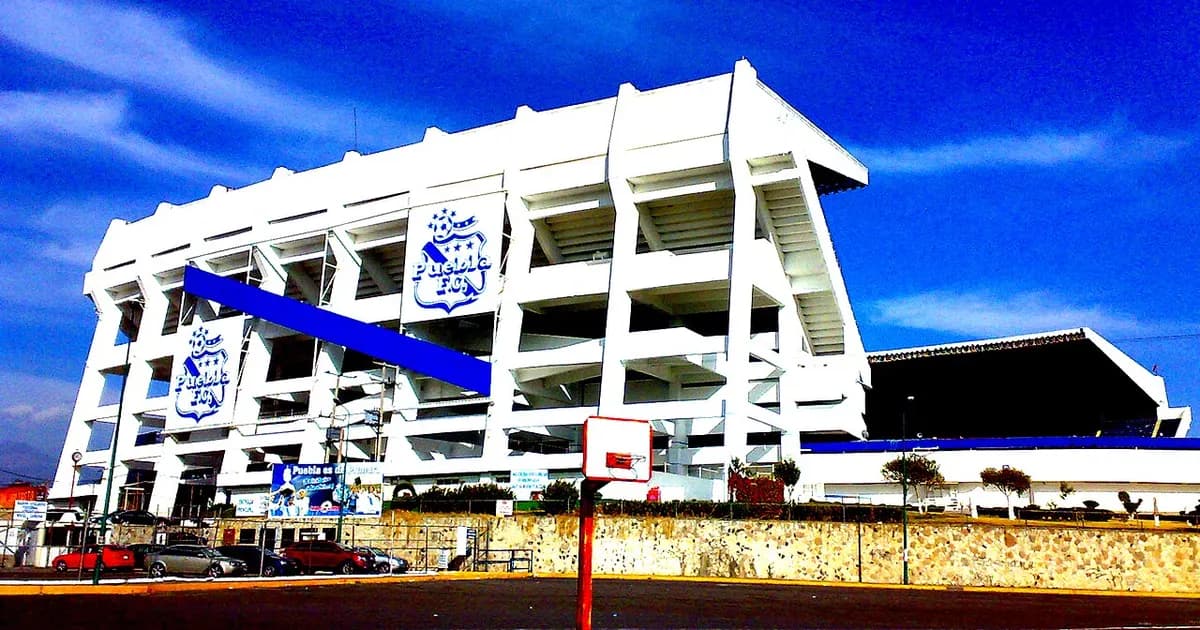 General view of a football stadium's pitch and stands before kickoff, with empty seats and green grass under stadium lights.